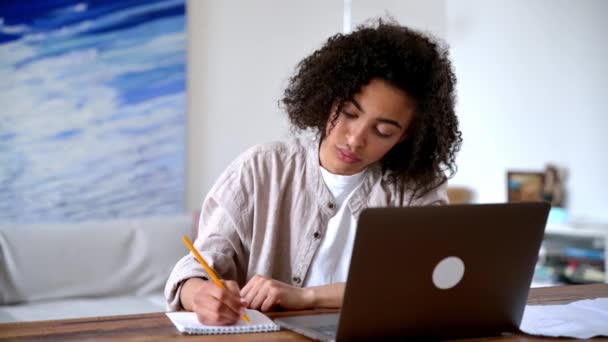 Ładna, odnosząca sukcesy młoda Afroamerykanka w stylowych ciuchach, freelancer lub student pracujący lub studiujący zdalnie, siedząca w salonie na laptopie, patrząca na ekran, robiąca notatki — Wideo stockowe