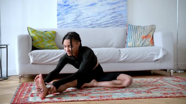 Sportliches und flexibles hübsches afroamerikanisches Mädchen in Sportbekleidung, engagiert sich für Fitness, führt einen gesunden Lebensstil, macht Stretching zu Hause auf dem Boden, schaut zur Seite, lächelt glücklich — Stockvideo