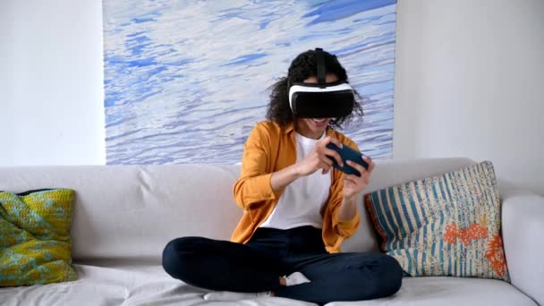 Cyberworld. Alegre alegre rizado joven afroamericana mujer sentada en el sofá con ropa casual, el uso de teléfonos inteligentes y la prueba de dispositivo de realidad virtual en el mundo digital, sonriendo felizmente — Vídeo de stock