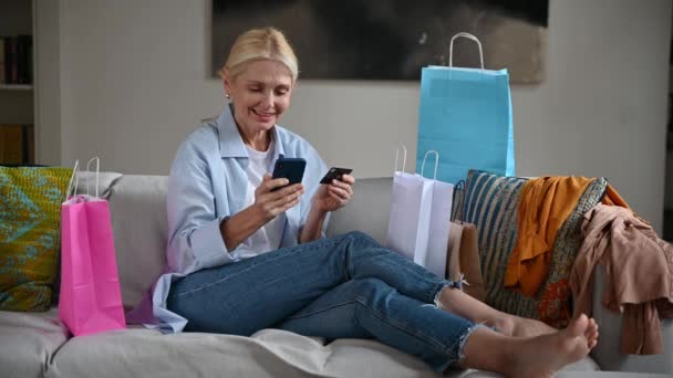 Compras en línea. Caucásico rubio elegante mujer de mediana edad adicta a las compras, sentado en el sofá en la sala de estar, utiliza el teléfono inteligente y la tarjeta de crédito para el pago en línea, hace un pedido en Internet, sonriendo — Vídeos de Stock