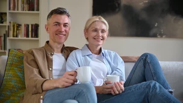 Feliz pareja casada caucásica adulta positiva con ropa casual sentada en un cómodo sofá en la sala de estar, sosteniendo tazas de té, mirando a la cámara, sonriendo. Marido y esposa están descansando en casa — Vídeos de Stock