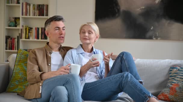 Houden van gelukkig volwassen getrouwd Kaukasisch paar in stijlvolle casual kleding, gelukkig man en vrouw zitten op de comfortabele bank in de woonkamer, het houden van kopjes thee, chatten, tijd thuis doorbrengen — Stockvideo