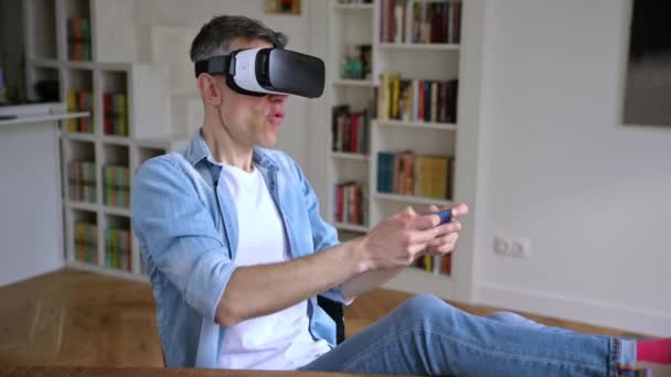 Захоплений чоловік середнього віку, який сидить за столом і жестикулює руками під час тестування нового додатка через VR-пристрій. Веселий зрілий чоловік використовує VR окуляри і тримає смартфон, концепція віртуальної реальності — стокове відео
