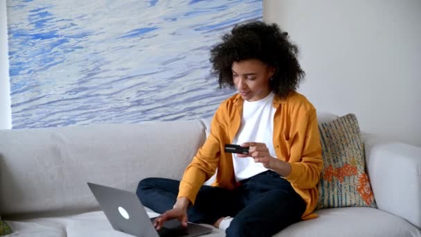 Online αγορές. Νεαρή όμορφη Αφροαμερικανή γυναίκα σε casual κομψά ρούχα κάθεται στον καναπέ, κάνει αγορές μέσω του Διαδικτύου, συμπληρώνει τα στοιχεία της πιστωτικής κάρτας για να πληρώσει για αγαθά ή κατ 'οίκον παράδοση — Αρχείο Βίντεο