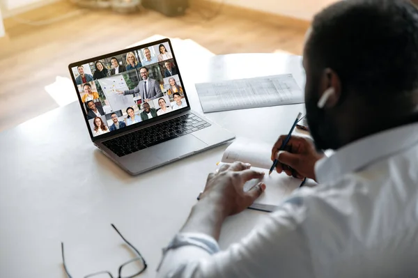 Online business seminar. View over the shoulder of an african american guy taking notes in a notebook during an online financial lecture, on a laptop screen are multiracial business colleagues — Stock Fotó