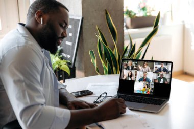 E-öğrenme. Odaklanmış Afrikalı Amerikalı adam evde masada oturuyor, dizüstü bilgisayarından online bir ders dinliyor, notlar alıyor, ekranda bir bayan öğretmen beyaz tahtada bilgi gösteriyor, ve diğer öğrenciler