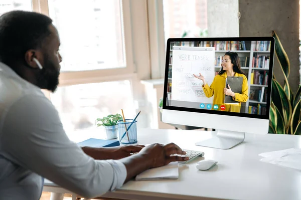 E-learning, online lesson. African American smart guy sits at home at work desk, uses computer, listening to an online lecture, female teacher on the screen shows information on a whiteboard — Photo