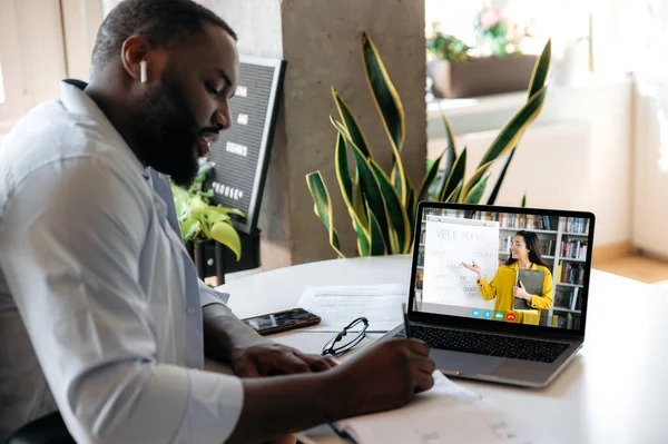 Online education. Focused african american guy sits at the table at home, listens to an online lesson by laptop, makes notes, on the screen female teacher shows information on a whiteboard, video call — Fotografia de Stock