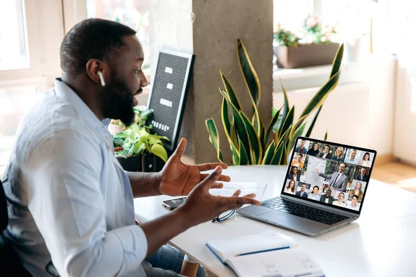 Virtual online meeting by video call. African American man communicating via video conference with multinational colleagues, gesturing with his hands, while sitting at home at work place, distant work — Stock Fotó