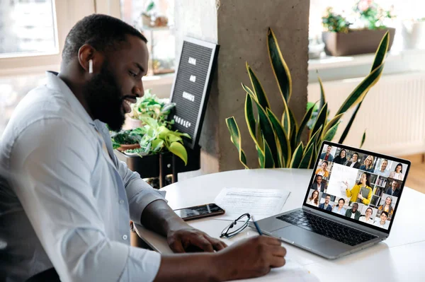 Online virtual meeting, e-learning. Focused african american man, student, sits at workplace, makes notes during online lesson, uses laptop, female teacher and students are on a screen — Fotografia de Stock
