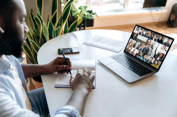 E-learning concept, online education. Focused african american guy taking notes in a notebook during an online business lecture, on a laptop screen is male mentor and diverse people, video call — Stock Fotó