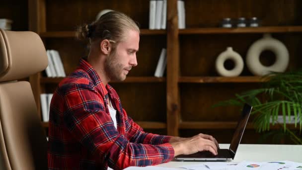 Inteligente atraente jovem adulto bem sucedido branco gerente ou freelancer com barba senta-se na mesa de trabalho, em camisa casual, usando um laptop, olhando através de documentos de negócios e gráficos, analisando — Vídeo de Stock