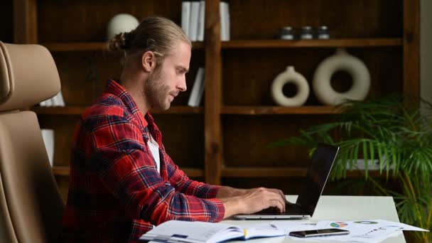 Успешный молодой кавказский фрилансер или менеджер с бородой, сидящий за столом в стильной рубашке, пользующийся ноутбуком, переписывающийся с друзьями или коллегами, думающий о проекте — стоковое видео