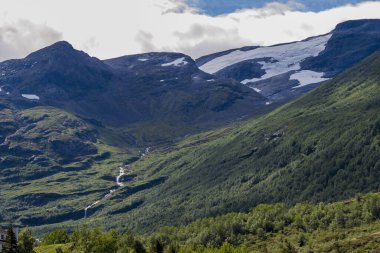 Tepesinde kar tarlaları olan büyük yeşil dağlar ve vadiye doğru inen bir dere.