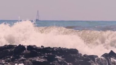 Denizin dalgaları kayaların üzerinde beyaz köpük oluştururken, bir yelkenli dipte yol alır.