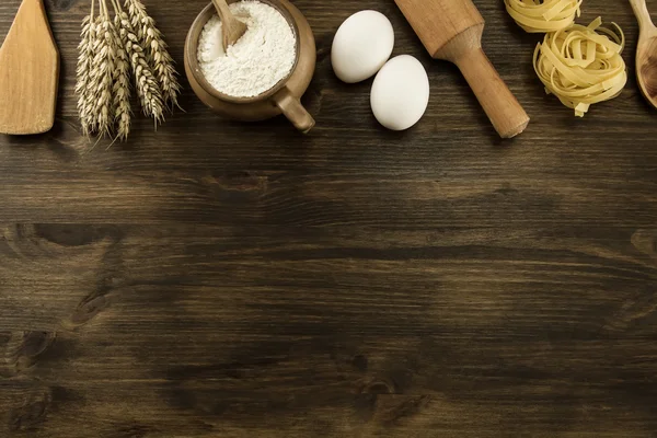 Pot of flour, wheat ears, pasta, eggs, kitchen utensils on wooden background. homemade, menu, recipe, mock up — Stock Photo, Image