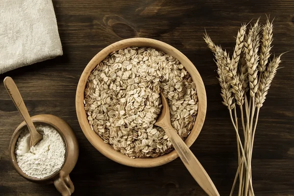 Copos de avena en un tazón de madera con una cuchara, espigas de trigo, olla de harina sobre la mesa. casero, menú, receta, maqueta — Foto de Stock