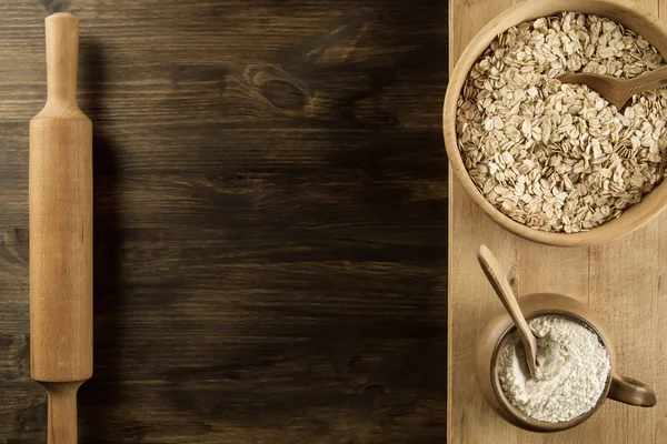 Copos de avena en un tazón de madera con una cuchara, espigas de trigo, olla de harina, utensilios de cocina en la mesa. casero, menú, receta, maqueta . — Foto de Stock