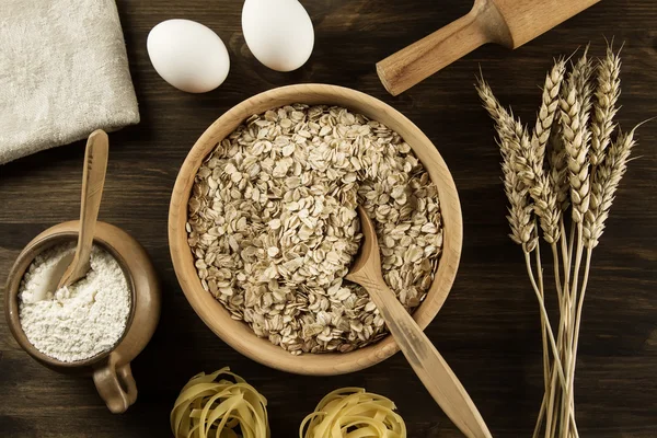 oatmeal flakes in a wooden bowl with a spoon, ears of wheat, pot of flour, kitchen utensils on the table. homemade, menu, recipe, mock up.