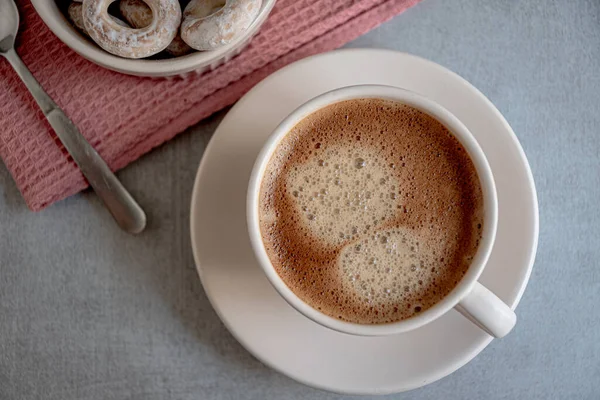 A cup of coffee on a saucer, bagels in a ceramic dish, a karalov-colored kitchen napkin on a gray background.