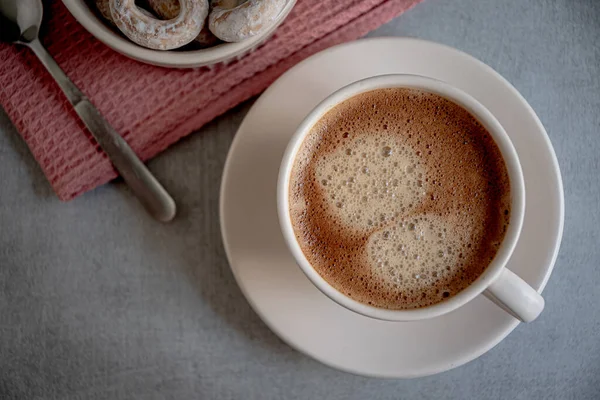 A cup of coffee on a saucer, bagels in a ceramic dish, a karalov-colored kitchen napkin on a gray background.