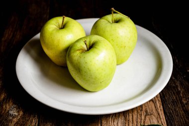 Üç yeşil elma beyaz bir seramik tabakta, ahşap bir masada..
