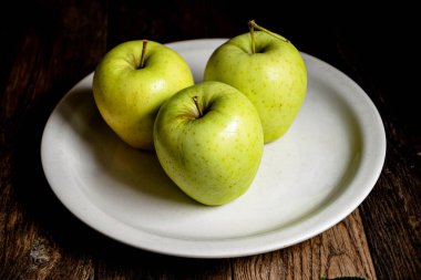 Üç yeşil elma beyaz bir seramik tabakta, ahşap bir masada..