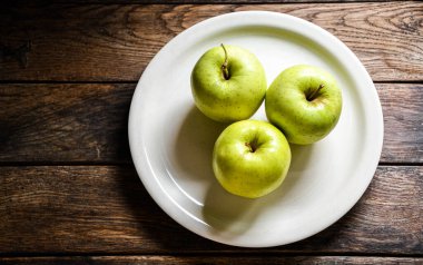Üç yeşil elma beyaz bir seramik tabakta, ahşap bir masada..