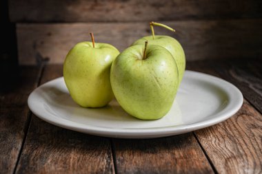 Üç yeşil elma beyaz bir seramik tabakta, ahşap bir masada..