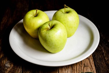 Üç yeşil elma beyaz bir seramik tabakta, ahşap bir masada..