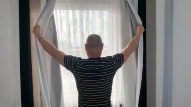 Kel adam pencerenin önünde duruyor, perdeleri açıyor, sokağa bakıyor, iş seyahati konsepti, turizm, yalnızlık, evde kalmak, karantina