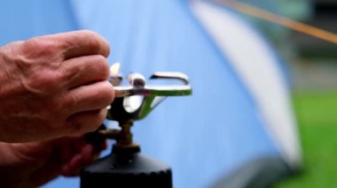 Turist kahvaltıda su kaynatıyor. Turist çaydanlığında. Kamp alanında gaz ocağında. Arka planda mavi bir çadır.