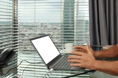 Adam bir dizüstü bilgisayarın arkasında çalışıyor. Modern Avrupa şehrinin penceresinin önündeki bir ofiste boş ekranla. İş fikirleri konsepti, işyeri.