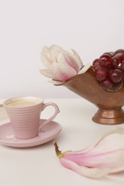 Beyaz bir masada bir fincan kapuçino, çiçek açan manolya tomurcukları, pembe arka planda beyaz bir kapakta bir kitap, tasarımcı için bir boşluk, bir davetiye formu, bahar havası konsepti.