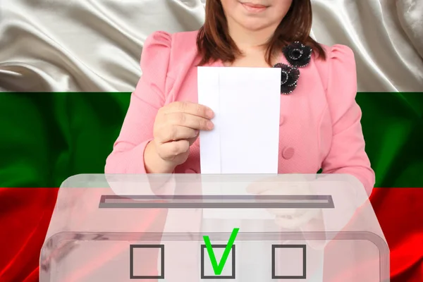 woman voter in a smart pink jacket lowers the ballot in a transparent ballot box against the background of the national flag of Bulgaria, the concept of state elections, referendum