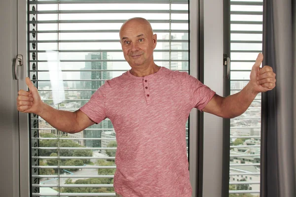 middle-aged bald man in a pink T-shirt stands by the window in the office, emotionally rejoices, smiles, the concept of success, achievement, lifestyle, holiday, good mood