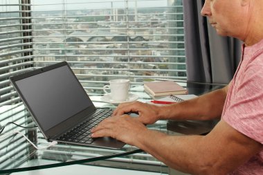 Adam bir dizüstü bilgisayarın arkasında çalışıyor. Modern Avrupa şehrinin penceresinin önündeki bir ofiste boş ekranla. İş fikirleri konsepti, işyeri.