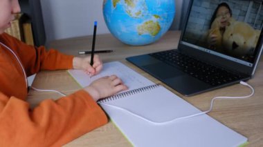 6-7 yaşlarındaki çocuk not defterine yazıp çiziyor, dizüstü bilgisayarın önünde ödev yapıyor, ekrandaki öğretmen internetten ders veriyor, uzaktan öğrenme kavramı, covid-19 'u kilitleme