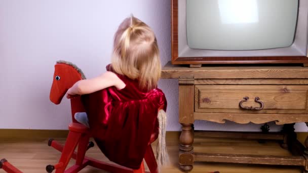 小さなブロンド女の子2歳で赤いベルベットのドレス座っているとスイングオンザ木製ヴィンテージロッキング馬でザ部屋の近くにザ 古いテレビ 空白の画面 子供時代の思い出コンセプト — ストック動画
