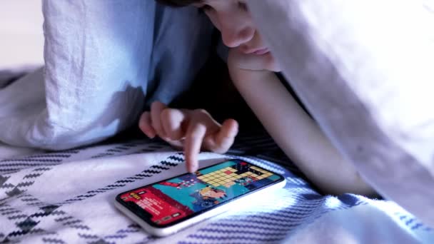 8歳の少年がベッドのカバーの下に横たわっていますスマートフォンのアプリケーションで遊び中毒不眠症の概念 — ストック動画