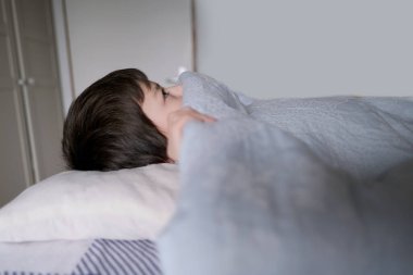 kid, a boy of 8 years old lies in bed, hiding under the covers, the concept of anxious sleep in schoolchildren, insomnia, night fears