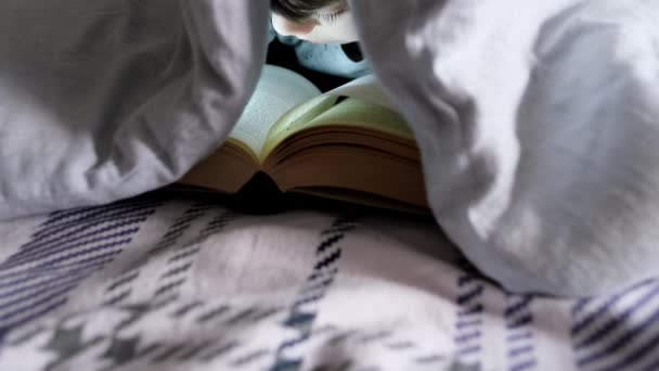 一个8岁的男孩躺在被窝里 躺在被窝里 阅读一本有趣的纸质书 上面有手电筒 知识概念 — 图库视频影像