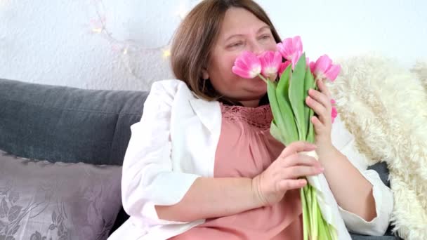 ピンクのチューリップの花束とソファの上に座っている大人の女性 それらを嗅ぎます くしゃみ 春の花粉アレルギーの概念 — ストック動画