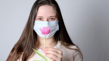 Genç bir kadın, uzun sarı saçlı, beyaz tıbbi maskeli bir kız yüzünün yanında pembe bir lale, bahar polen alerjisi konsepti, anneler günü, koronavirüs yüzünden koku kaybı.