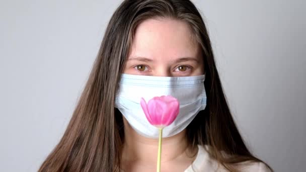 若い女性ブロンドの髪の長い白い医療マスクの女の子は彼女の顔の近くにピンクのチューリップを1つ持っています春の花粉アレルギーの概念 母の日 コロナウイルスによる匂いの損失 — ストック動画