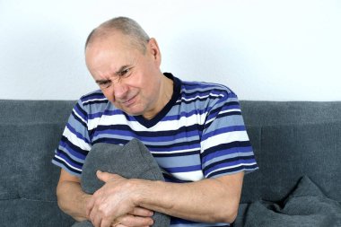 Yetişkin kel bir adam kanepeye oturur, elini hipokondriyumun sağ tarafında tutar, yüzünde bir acı ifadesi, karaciğer hastalığı konsepti, safra kesesi.