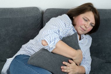 Orta yaşlı bir kadın kanepede oturuyor, yastığı karnına bastırıyor, acı çekmekle yüz yüze, mide ağrısı konsepti, mide iltihabı, mide bulantısı.