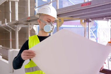 Genç erkek ustabaşı, mimar, inşaat alanındaki teftiş sırasında koruyucu solunum cihazı, kağıt üzerine proje planına bakıyor, değerlendirmeler yapıyor, yapıların izlenmesi kavramını inceliyor.