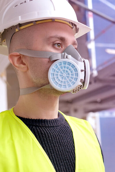 young male builder foreman, engineer or architect in white hardhat and protective respirator during inspection in construction site, concept of monitoring repair of buildings, structures