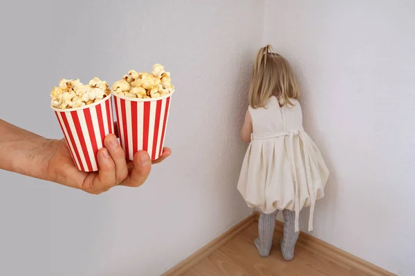 small child in a white dress, baby, a blonde girl stands in the corner of the room, hand holds out glasses of popcorn, concept of parenting, abuse, punishment